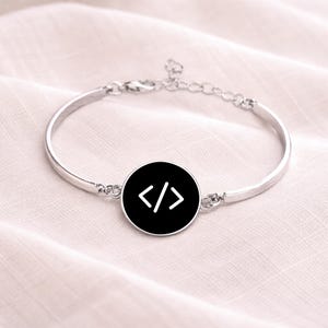 Peut inclure: Un bracelet jonc argenté avec un charm circulaire noir. Le charm présente un symbole de code blanc. Le bracelet est posé sur une surface douce et claire.