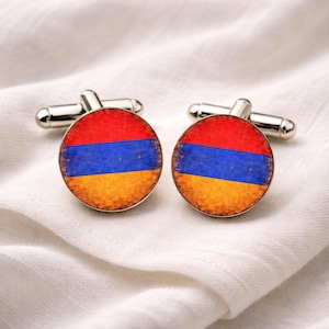 Peut inclure: Paire de boutons de manchette argentés avec un motif circulaire représentant le drapeau arménien. Le drapeau présente des bandes horizontales rouge, bleu et orange, avec un aspect vieilli. Les boutons de manchette sont posés sur un fond en tissu blanc.