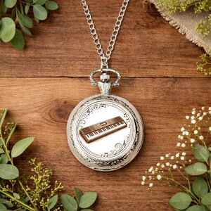 May include: Silver pocket watch pendant with a detailed keyboard design on the face. The watch hangs from a silver chain and features musical notes around the keyboard image. The watch is set against a wooden background.