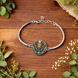 May include: A silver bangle bracelet with a circular pendant. The pendant showcases a detailed bear illustration with teal and gold accents. The bracelet is displayed on a wooden surface, with decorative foliage around it.