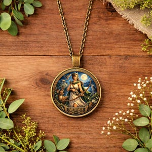 May include: A bronze pendant necklace with a circular pendant. The pendant features an illustration of Artemis, the Greek goddess of the hunt, with a bow and arrow, a deer, and a full moon. The word "ARTEMIS" is printed on the pendant.