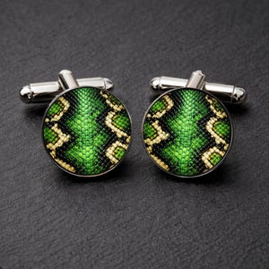 Puede incluir: Un par de gemelos plateados con un diseño redondo de piel de serpiente verde y amarilla. Los gemelos tienen un acabado metálico brillante y un diseño clásico, adecuados para ocasiones formales o para añadir un toque único.