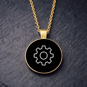May include: A gold necklace with a circular pendant. The pendant has a white gear symbol on a black background, set within a gold frame. The chain is gold. The necklace is shown against a dark, textured surface.