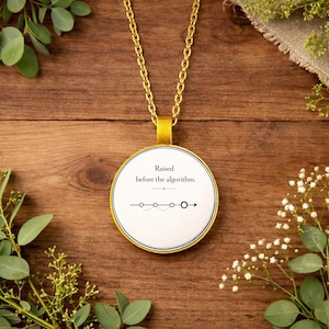 Op de afbeelding: Een gouden ketting met een ronde, witte hanger. De hanger heeft de tekst "Raised before the algorithm." en een lijn met cirkels en een pijl. De ketting ligt op een houten ondergrond met groen.