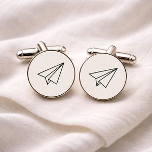 Puede incluir: Gemelos plateados con una cara circular blanca. Cada gemelo presenta un dibujo lineal negro de un avión de papel. Se muestran sobre una tela blanca texturizada.