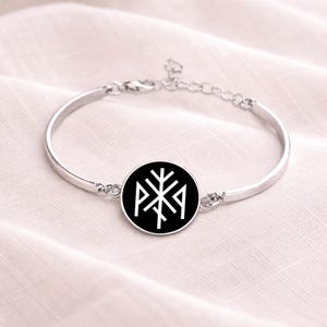 May include: A silver-toned bangle bracelet with a circular black pendant. The pendant features a white geometric design. The bracelet has an adjustable chain closure and is displayed on a soft, light-colored surface.