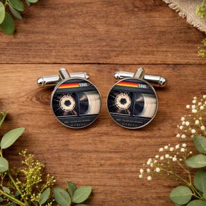 May include: A pair of silver cufflinks with a retro VHS tape design. Each cufflink features a detailed image of a cassette tape with the word "VHS" and a rainbow stripe. The cufflinks are set against a wooden background.