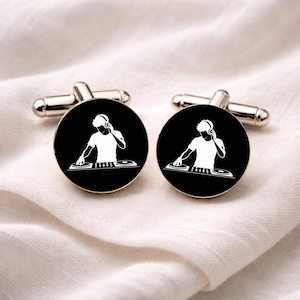 Könnte beinhalten: Paar silberne Manschettenknöpfe mit schwarzem, kreisförmigem Design. Jeder Manschettenknopf zeigt eine weiße Silhouette eines DJs mit Kopfhörern und Plattenspielern. Die Manschettenknöpfe werden auf einem weißen Stoffhintergrund präsentiert.