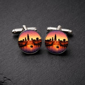 May include: A pair of silver cufflinks with a round design featuring a cityscape at sunset. The image shows a skyline with tall buildings, a river, and a vibrant orange and purple sky. The cufflinks have a classic, elegant design.