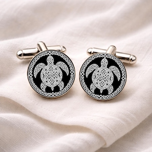 May include: A pair of silver cufflinks with a black circular face. Each cufflink features a white turtle design with intricate Celtic knotwork. The cufflinks are displayed on a white fabric background.
