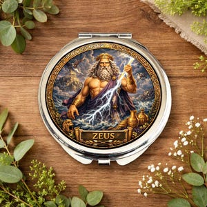Könnte beinhalten: Ein silberner Kompaktspiegel mit einer Illustration von Zeus, dem griechischen Gott des Donners, der einen Blitz hält. Das Bild ist mit einem goldenen griechischen Schlüsseldesign umrahmt und der Name "ZEUS" ist darunter abgebildet.