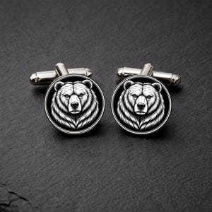 Puede incluir: Gemelos plateados con un diseño de cabeza de oso en blanco y negro. El rostro del oso está detallado con pelaje. Los gemelos tienen forma redonda y un diseño clásico.