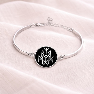 May include: Silver-toned bangle bracelet with a circular black charm featuring a white geometric design. The bracelet has a simple, elegant design and is likely made of metal. The charm is the focal point, with its contrasting colors and intricate pattern.