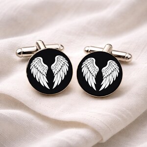 May include: Silver cufflinks with a black circular face, each displaying a white wing design. The cufflinks are presented on a white, textured fabric.