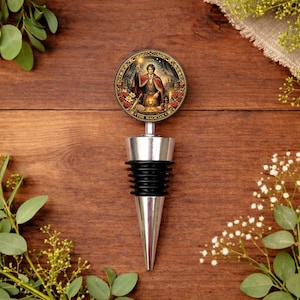 May include: A wine bottle stopper with a silver and black design. The circular emblem features an illustration of a figure in a red cloak, holding a staff and a chalice. The text "THE MAGICIAN" is printed on the emblem.