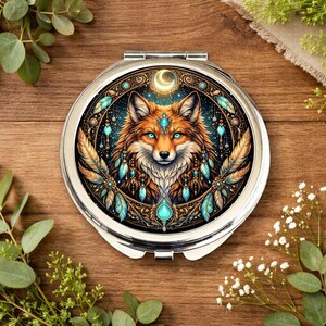May include: A silver compact mirror featuring a detailed illustration of a fox with blue eyes, decorated with feathers and turquoise accents. The design includes a crescent moon and celestial elements, set against a dark background.