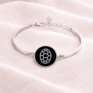 Könnte beinhalten: Ein silberfarbenes Armband mit einem kreisförmigen, schwarzen Anhänger mit einem weißen geometrischen Design. Das Armband hat einen verstellbaren Kettenverschluss. Das Design des Anhängers ähnelt einem Schildkrötenpanzer.