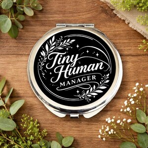 Può includere: Un specchietto compatto rotondo e argentato con una superficie nera. La superficie dello specchio presenta il testo bianco "Tiny Human Manager" in un carattere decorativo, circondato da elementi floreali bianchi. Lo specchio è posto su uno sfondo di legno con vegetazione.