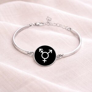 Könnte beinhalten: Silberfarbenes Armreif-Armband mit einem schwarzen, runden Anhänger, der ein weißes Transgender-Symbol zeigt. Das Armband hat einen verstellbaren Kettenverschluss und liegt auf weichem, hellem Stoff.