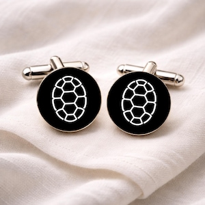 May include: A pair of cufflinks with a black background and a white geometric design. The cufflinks have a silver-colored metal backing and are displayed on a white fabric surface. The design is an oval shape made of connected hexagons.