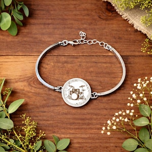 Op de afbeelding: Zilverkleurige armband met een ronde bedel met een drumstelontwerp. De bedel heeft muzieknoten rond de rand. De armband ligt op een houten oppervlak met groen.