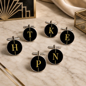 Op de afbeelding: Een set van zes ronde, zilverkleurige manchetknopen met een zwart oppervlak en een enkele gouden letter in het midden. De letters zijn H, T, K, E, P en N. Elke manchetknop heeft een verticale gouden lijn die door het midden van de letter loopt.
