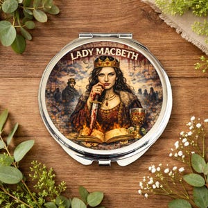 May include: A circular, silver compact mirror with an illustration of Lady Macbeth. The image shows a crowned woman holding a bloody dagger, with the words "LADY MACBETH" above the image.