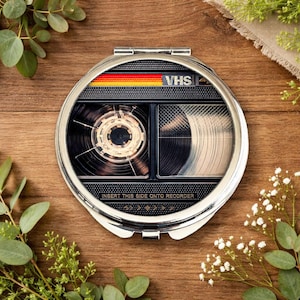 Könnte beinhalten: Runder, silberner Kompaktspiegel mit Retro-VHS-Kassetten-Design. Der Spiegel hat ein schwarz-silbernes Innere mit einem detaillierten Bild einer VHS-Kassette. Die Oberseite hat einen rot-gelb-orangen Streifen mit dem Wort "VHS". Der Text "INSERT THIS SIDE ONTO RECORDER" befindet sich unten.