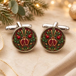 May include: A pair of circular silver cufflinks featuring a detailed enamel design of a split pomegranate with red seeds, surrounded by green leaves and stems against a deep red background with gold-toned geometric accents and a metallic finish.