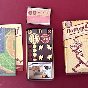 Op de afbeelding: Bordspel "Bottom of the 9th" met honkbalthema. De doos toont een honkbalspeler. Het spel bevat dobbelstenen, fiches, kaarten en een bord, alles op een bordeauxrode ondergrond.