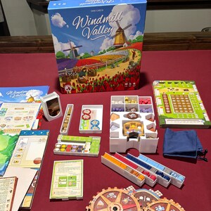 May include: A board game titled "Windmill Valley" with a box featuring a scenic illustration of windmills and flower fields. The game includes various components like game boards, tokens, and wooden gears, all arranged on a red surface.