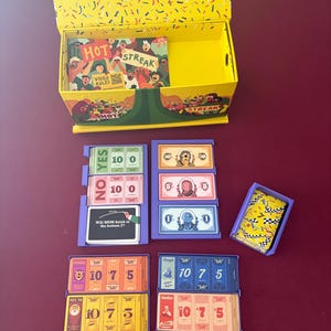 May include: A board game set with a yellow box and various game pieces. The box lid is open, revealing a card deck with the words "HOT STREAK" and "VIDEO RULES". Several game cards and a small container with a yellow and black pattern are visible.