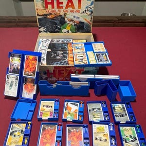 May include: A board game titled "HEAT Pedal to the Metal" with a box featuring a race car illustration. The game includes blue plastic trays, cards, and miniature race car pieces. The game is set on a red surface.