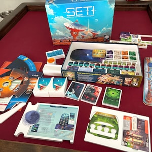 May include: A board game titled "SETI" with a box featuring a space-themed illustration. Game components include cards, tokens, and a game board, all arranged on a red surface. The game box reads "Search for Extraterrestrial Intelligence."