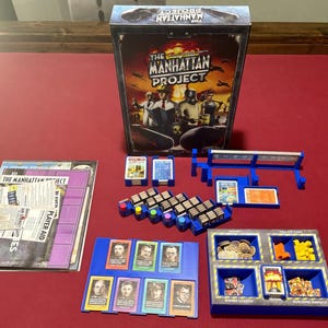 May include: The Manhattan Project board game box featuring a central illustration of scientists and military personnel. Includes a game board, player aid cards, blue plastic card holders, small wooden resource tokens, and a blue organizer tray.