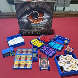 May include: The board game "RECALL" is displayed, with a box showing an eye illustration. The game's components, including cards and tokens, are arranged in blue trays. The game is set on a red surface.