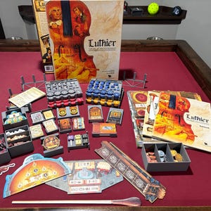 Puede incluir: Juego de mesa "Luthier: The Art of the Instrument" con componentes sobre una superficie roja. La caja muestra una ilustración de violín. Incluye cartas, fichas y piezas de juego.