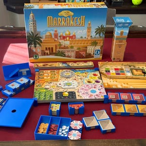 May include: The board game "Marrakech" is displayed with its components: a game board, cards, and wooden tokens. The game is set on a red surface, with a box featuring a city illustration.