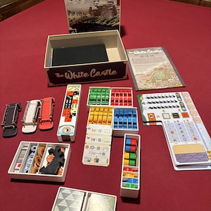 May include: A board game titled "The White Castle" with various game pieces arranged on a red surface. The game includes a box with artwork of a castle, dice, cards, and colorful game tokens in white containers.