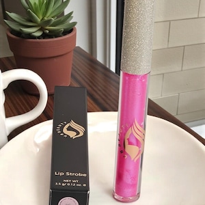 Può includere: Un lucidalabbra rosa e la sua scatola nera sono esposti su un piatto bianco. Il lucidalabbra ha un tappo argentato glitterato e un tubetto rosa. La scatola riporta "Lip Strobe" e "06". Una piccola pianta grassa in vaso è sullo sfondo.