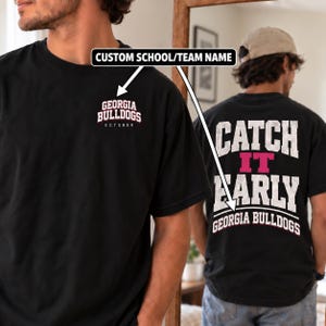 May include: A black t-shirt featuring white and pink text. The front left chest displays "GEORGIA BULLDOGS OCTOBER". The back features large white text reading "CATCH IT EARLY" with "GEORGIA BULLDOGS" printed below in white and pink lettering.