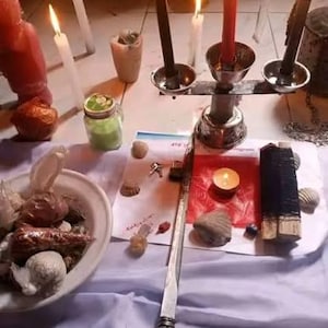 Puede incluir: Un candelabro de metal con dos velas negras y una vela roja, rodeado de varios objetos, incluyendo un cuenco blanco lleno de conchas, un frasco verde, una varilla de metal larga, un peque&ntilde;o cuadrado de papel rojo y varias velas blancas encendidas.