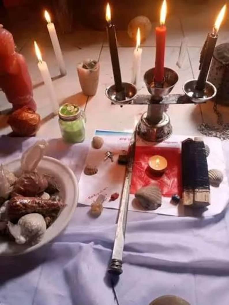 Puede incluir: Un candelabro de metal con dos velas negras y una vela roja, rodeado de varios objetos, incluyendo un cuenco blanco lleno de conchas, un frasco verde, una varilla de metal larga, un peque&ntilde;o cuadrado de papel rojo y varias velas blancas encendidas.