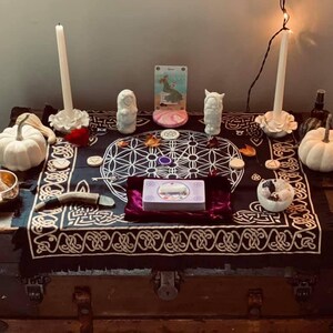 Puede incluir: Un mantel de altar negro con un patr&oacute;n geom&eacute;trico blanco de flor de la vida y bordes de nudos celtas, que incluye dos velas blancas, dos calabazas blancas, un cuenco tibetano de lat&oacute;n, una baraja de tarot, peque&ntilde;as figuras de piedra y cristales.