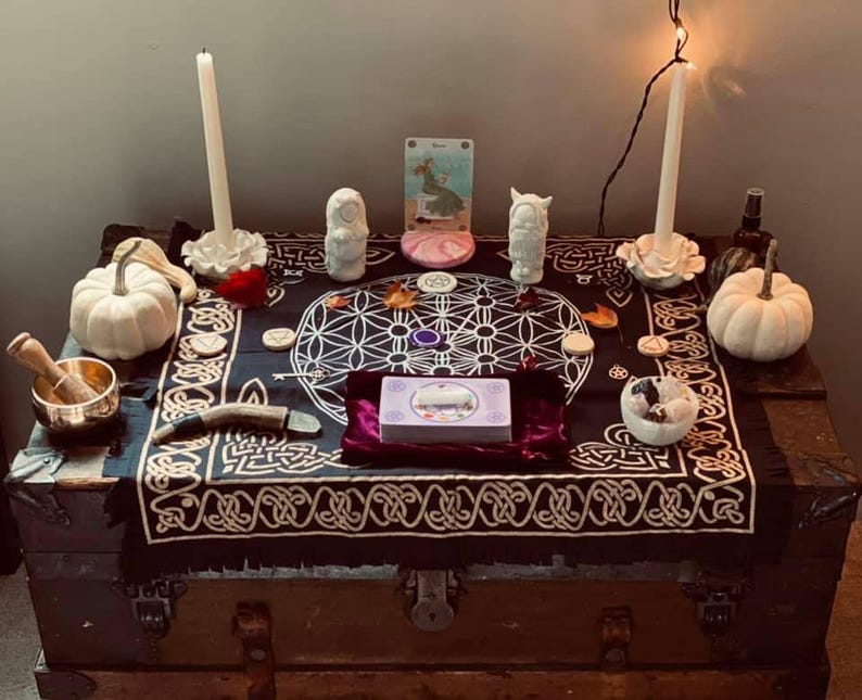 Puede incluir: Un mantel de altar negro con un patr&oacute;n geom&eacute;trico blanco de flor de la vida y bordes de nudos celtas, que incluye dos velas blancas, dos calabazas blancas, un cuenco tibetano de lat&oacute;n, una baraja de tarot, peque&ntilde;as figuras de piedra y cristales.