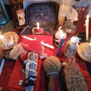 Puede incluir: Una colecci&oacute;n de objetos rituales que incluye una tela roja, varias botellas de vidrio, velas blancas y moradas, una maraca de madera con red, un manojo de fibras secas y un peque&ntilde;o estuche abierto con un estampado oscuro sobre una base roja.