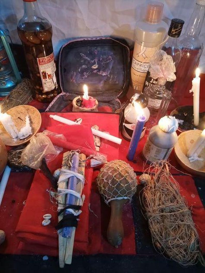 May include: A collection of ritual items including a red cloth, several glass bottles, white and purple candles, a wooden maraca with a net covering, a bundle of dried fibers, and a small open case with a dark graphic print on a red surface.