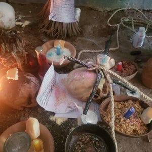 Pu&ograve; includere: Una collezione di oggetti rituali che include diverse ciotole di terracotta riempite di cereali, piccole candele blu, gialle e rosse, un teschio avvolto in una corda spessa, una bottiglia di vetro con liquido rosso e fogli scritti a mano.