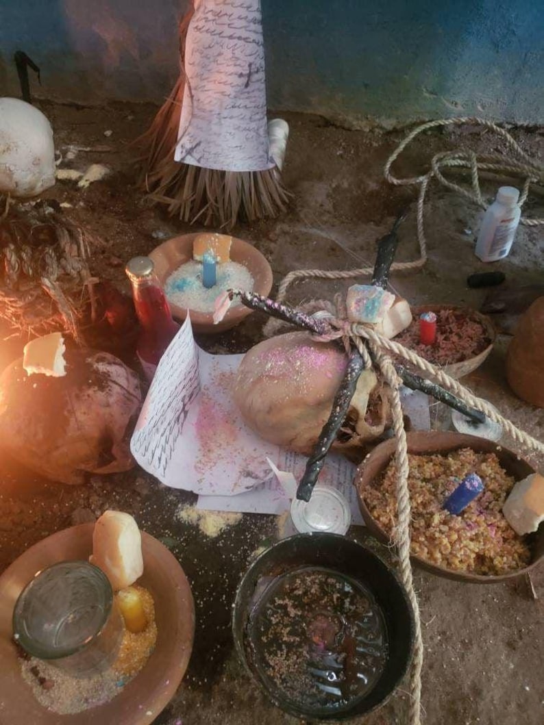Pu&ograve; includere: Una collezione di oggetti rituali che include diverse ciotole di terracotta riempite di cereali, piccole candele blu, gialle e rosse, un teschio avvolto in una corda spessa, una bottiglia di vetro con liquido rosso e fogli scritti a mano.