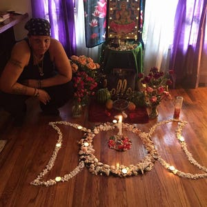 May include: A floor arrangement of white seashells and small stones forming a triple moon symbol on a wooden floor. A central white pillar candle sits within the circle, surrounded by smaller tea light candles placed along the shell outline.
